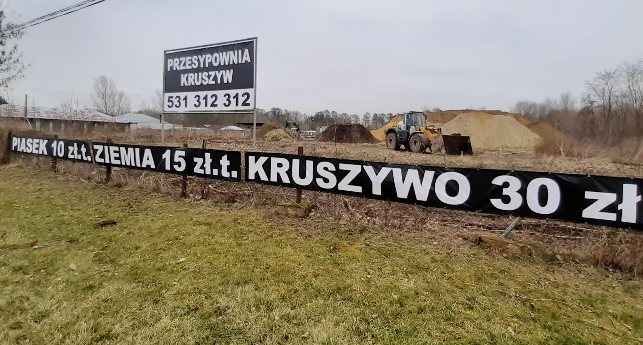 Plac z hałdami piasku, ziemi i kruszywa, widoczny baner reklamowy z cenami, w tle pracuje ładowarka kołowa.
