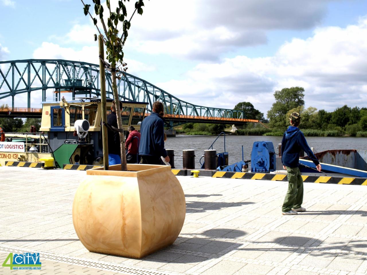 Duża, jasna donica miejska z młodym drzewkiem na tle rzeki, mostu kratownicowego i zacumowanej barki.