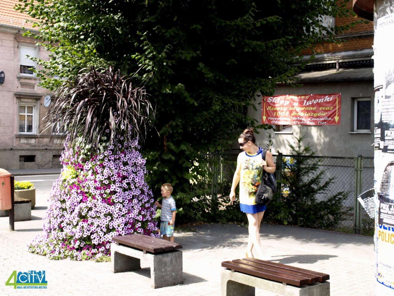 Wieża kwiatowa z fioletowych petunii i ciemnych traw ozdobnych na placu miejskim, obok betonowej ławki i przechodzących osób.