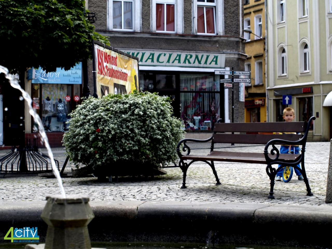 Miejski plac z fontanną, ławeczką i małym chłopcem na rowerze biegowym, w tle budynki z szyldami sklepów i drogowskazami.