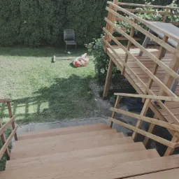 Nowa drewniana konstrukcja tarasu z schodami w ogrodzie, widok z góry na świeżo zbudowany taras z jasnego drewna, na trawie krzesło i narzędzia.