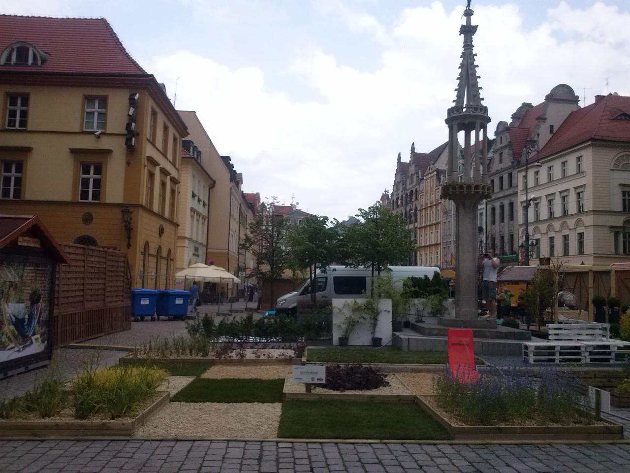 Kwadratowy ogródek pokazowy z trawą i kamieniami na brukowanym placu miejskim, z zabytkową kolumną i budynkami w tle.