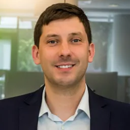 Portret uśmiechniętego mężczyzny w ciemnej marynarce i jasnej koszuli na tle biurowym z kolumną i oknami.