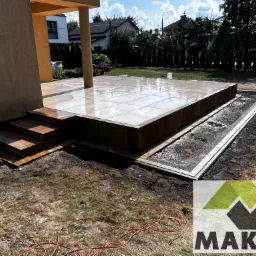 Nowoczesny taras z jasnych płytek ceramicznych, otoczony obrzeżem z kostki. Widoczne schody i przygotowane miejsce pod nasadzenia. W tle budynek mieszkalny i zieleń.