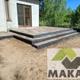 Makar - Ułożone schody z kostki brukowej prowadzące do drzwi tarasowych domu. Czarna obwódka kostki. Widoczne drzewa w tle. Logo firmy w prawym dolnym rogu.