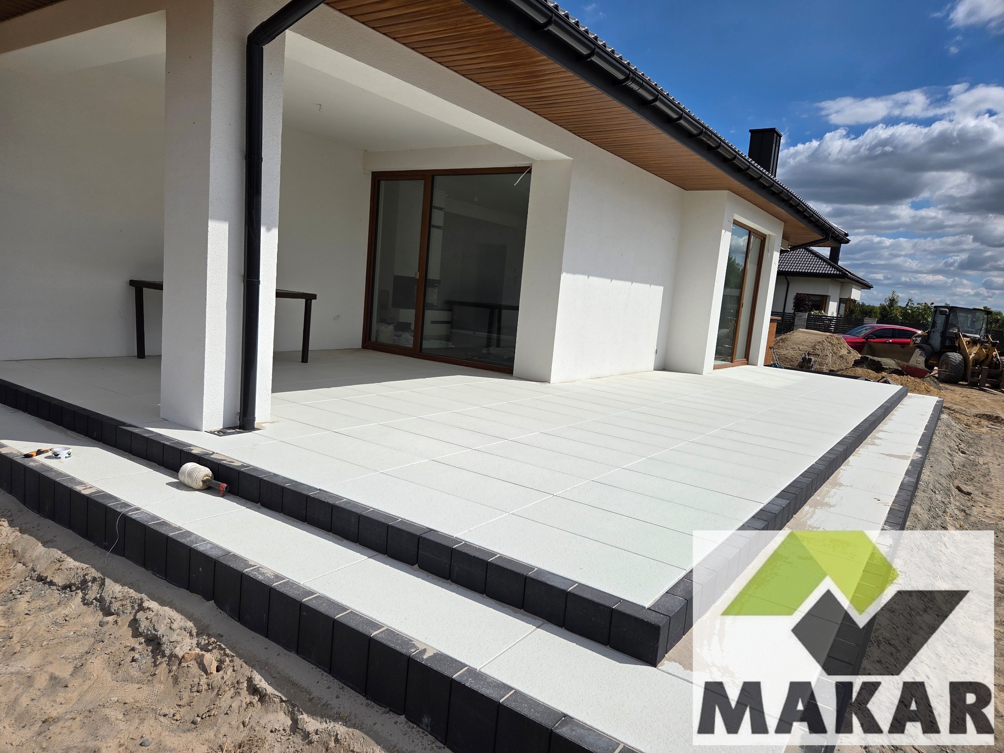 Nowoczesny taras z jasnych płyt, obrzeża z ciemnej kostki. Widoczne schody wejściowe i fragment ogrodu z koparką w tle.  Słoneczny dzień.