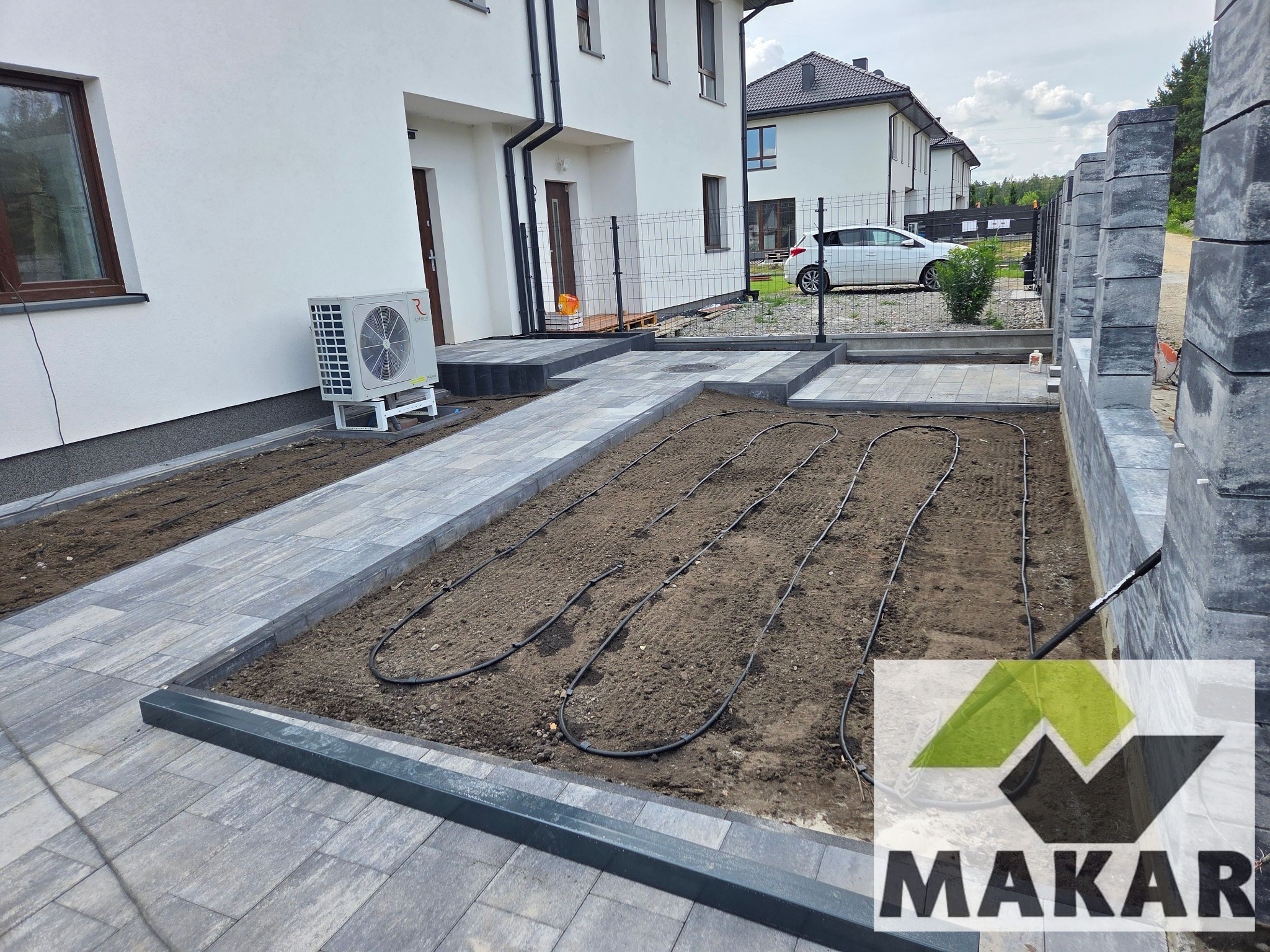 Nowoczesna posesja z ułożoną kostką brukową i systemem nawadniania w gruncie. Widoczny fragment ogrodzenia z betonowych bloczków i klimatyzator na elewacji budynku.