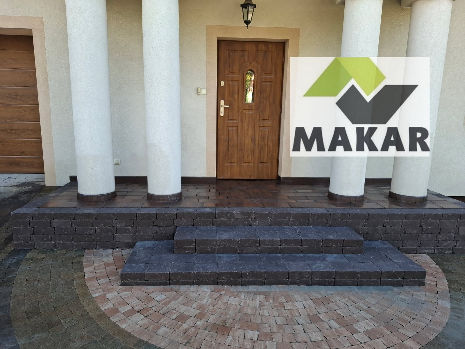 Eleganckie schody wejściowe z ciemnej kostki brukowej, prowadzące do drewnianych drzwi frontowych. Kolista kostka brukowa w odcieniach brązu i beżu.