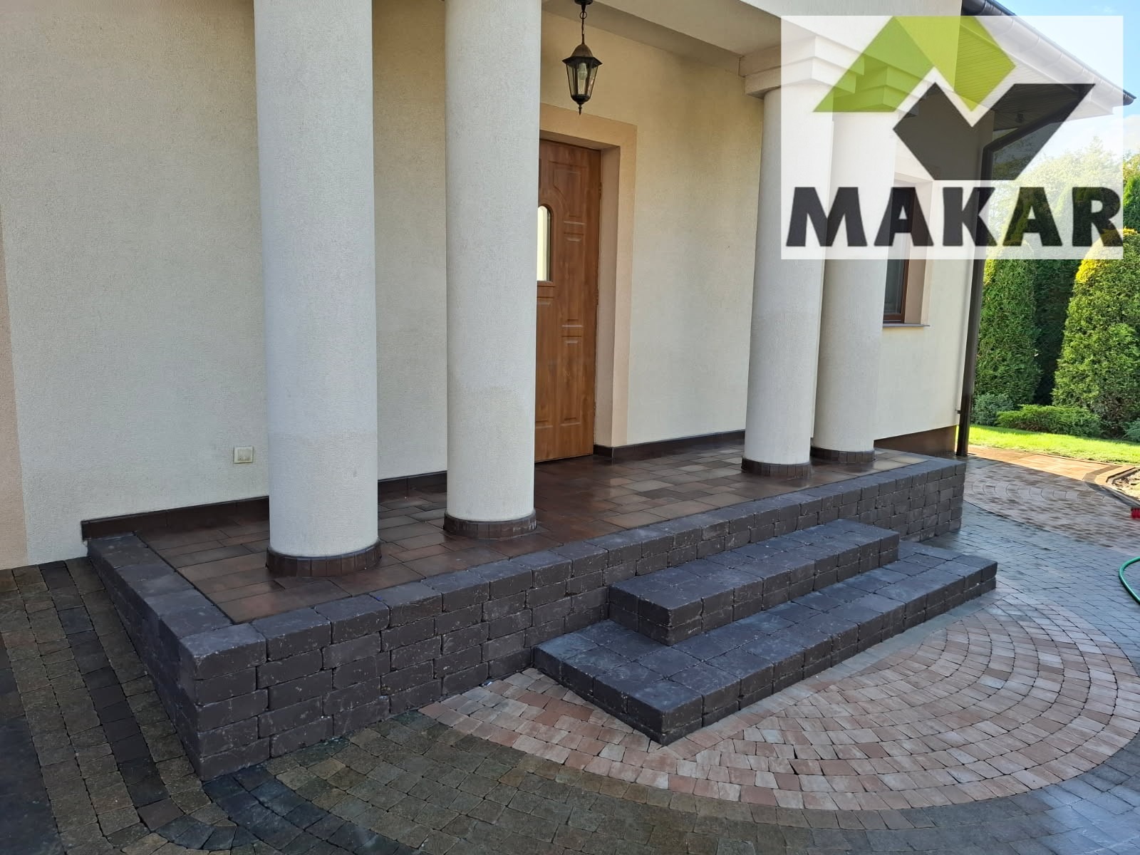 Eleganckie schody wejściowe z ciemnej kostki brukowej prowadzą do domu z kolumnami. Okrągły wzór z kostki na podjeździe. Widoczny fragment elewacji i drzwi.