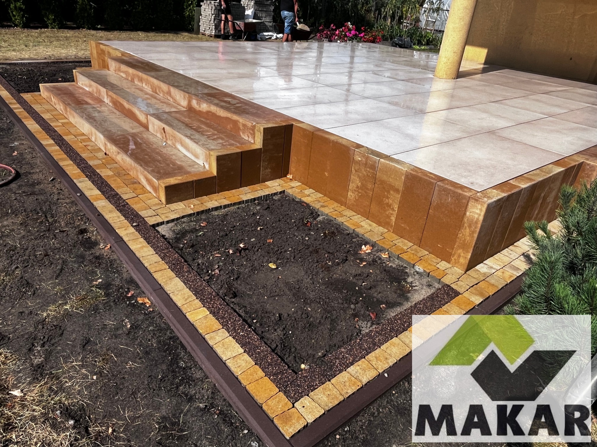 Wykończony taras z kamiennymi schodami i rabatą kwiatową obrzeżoną kostką, z widocznym logo firmy w prawym dolnym rogu zdjęcia.