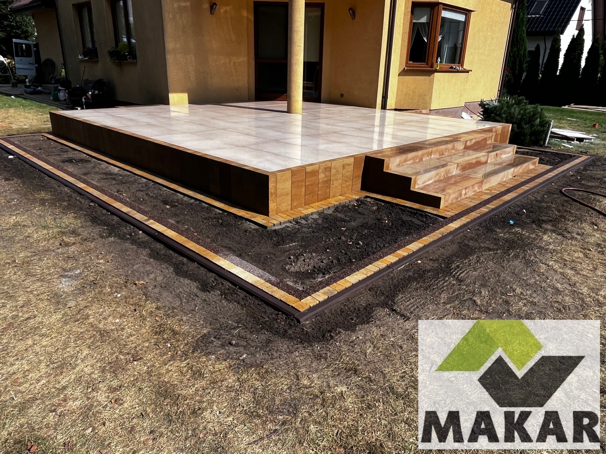 Wykończony taras z jasnych płytek, z niskimi schodami i obramowaniem z kostki, przylegający do budynku z kolumną. W tle trawnik i inne budynki.