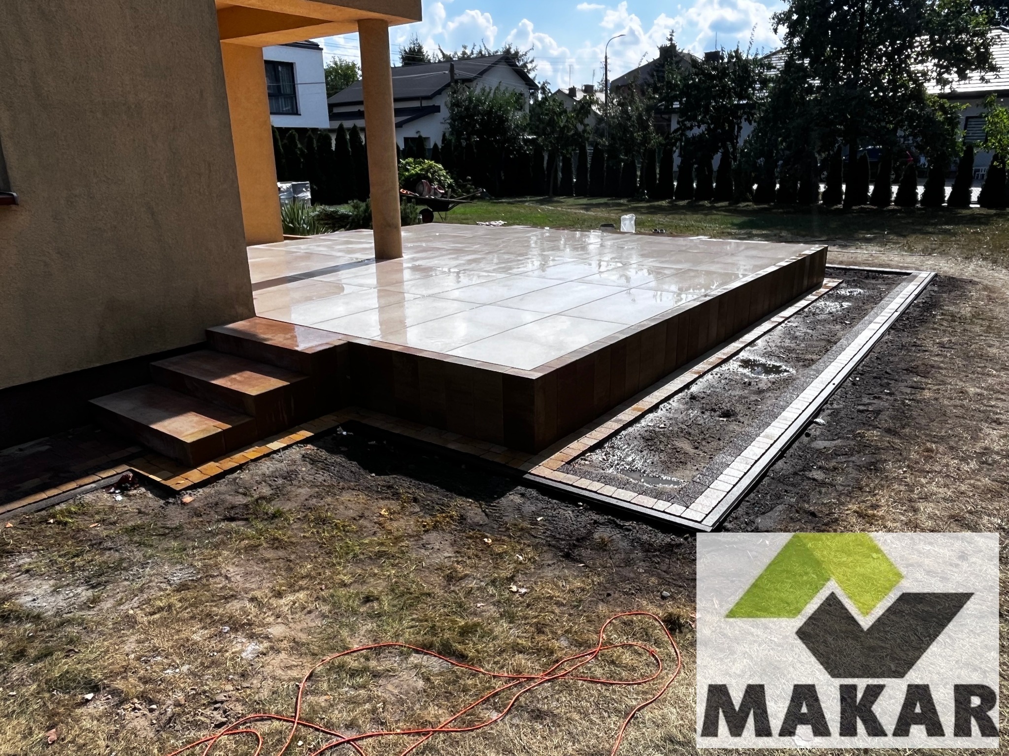 Nowoczesny taras z jasnych płytek ceramicznych, otoczony obrzeżem z kostki. Widoczne schody i przygotowane miejsce pod nasadzenia. W tle budynek mieszkalny i zieleń.