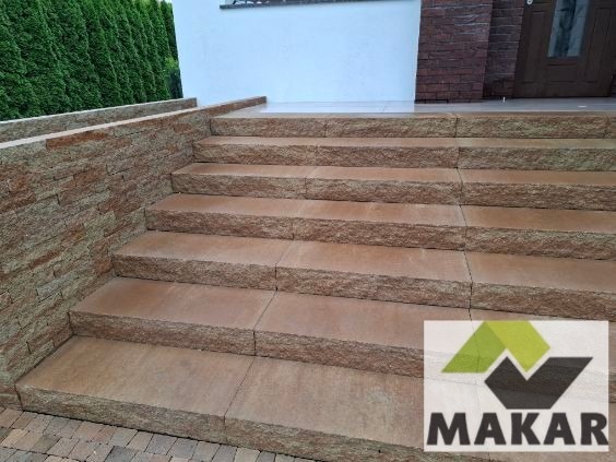 Wykończone schody zewnętrzne z kamienia naturalnego w odcieniach brązu, prowadzące do wejścia domu. Widoczna faktura kamienia i logo firmy w prawym dolnym rogu.