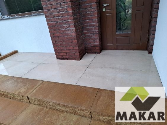 Wykończone płytkami wejście do domu z ceglanym filarem i brązowymi schodami. Widoczny fragment drzwi i logo firmy w rogu zdjęcia.