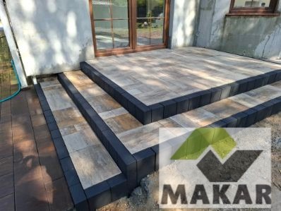 Wykończony taras z kostki brukowej z trzema stopniami i obramowaniem z ciemnej kostki. Widoczne drzwi balkonowe i fragment elewacji budynku.