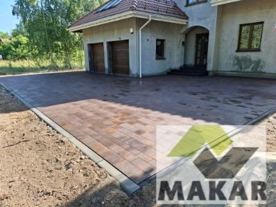 Ułożona, brązowa kostka brukowa na podjeździe przed domem z garażem. Widoczne obrzeża i częściowo niezabudowany teren wokół. Kostka ułożona w jodełkę.