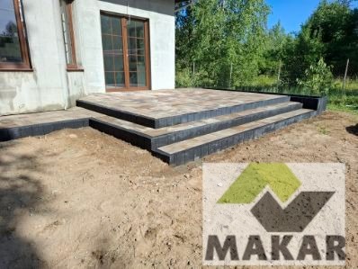 Ułożone schody z kostki brukowej prowadzące do drzwi tarasowych domu. Czarna obwódka kostki. Widoczne drzewa w tle. Logo firmy w prawym dolnym rogu.