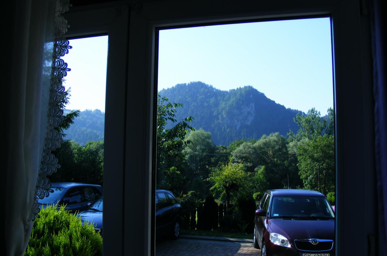 Widok z okna na górzysty krajobraz z zaparkowanymi samochodami na podjeździe, z fragmentem firanki z koronką po lewej stronie.