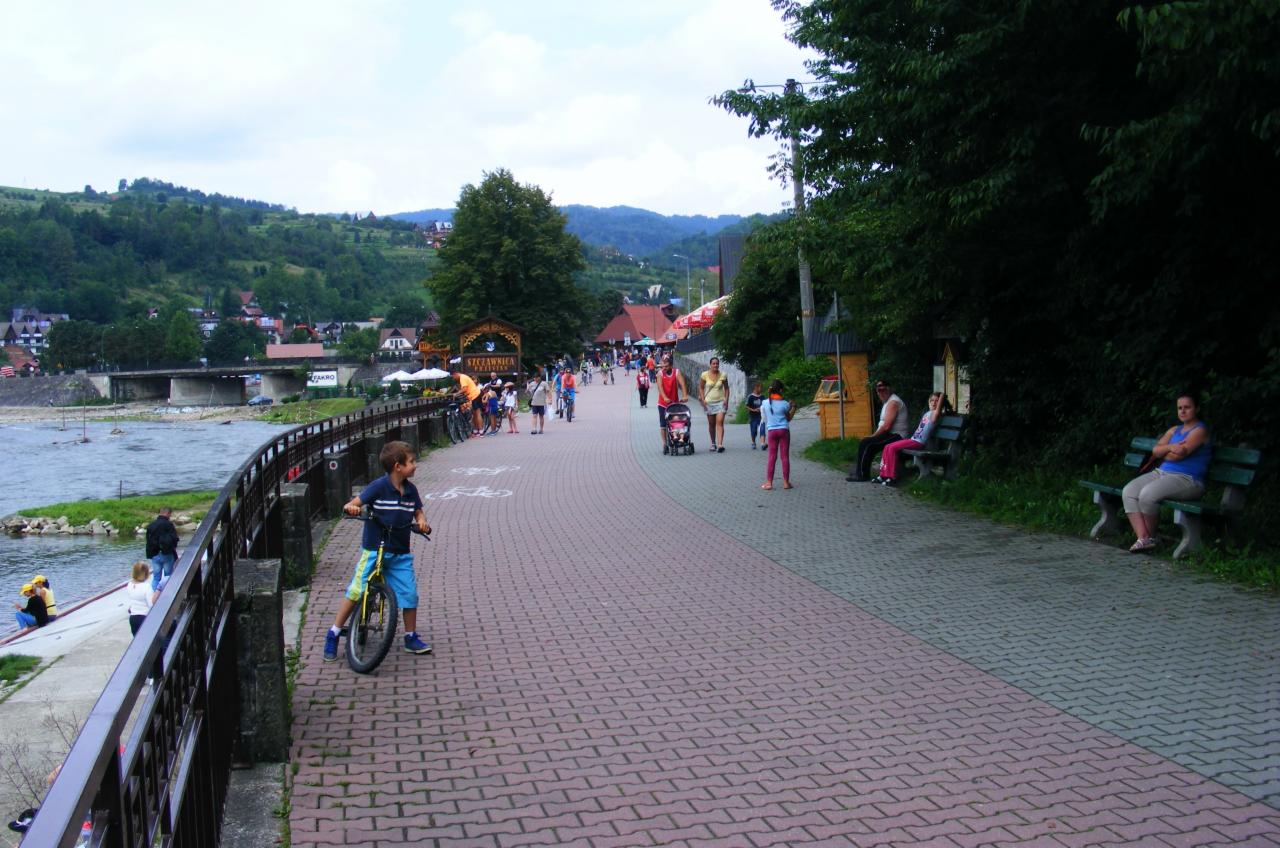 Ceglana ścieżka spacerowa w Szczawnicy, z widokiem na rzekę i góry, gdzie chłopiec jedzie na rowerze, a w tle widać spacerujących ludzi.