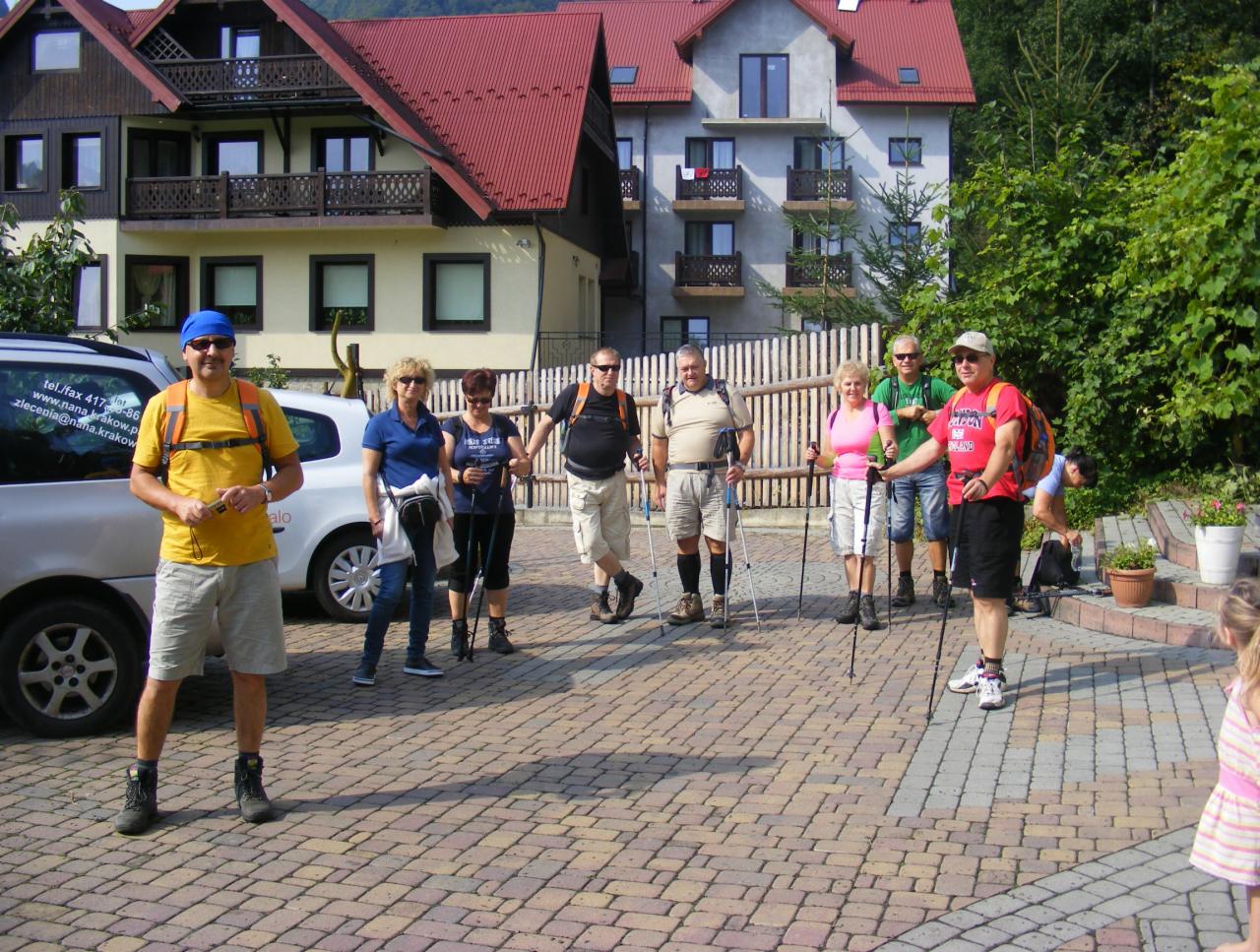 Grupa turystów z kijkami na brukowanym placu przed budynkami w górskim kurorcie.