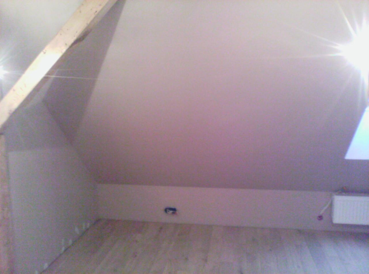 Poddasze z ukośnym dachem, ściany wykończone płytami, widoczne drewniane belki stropowe i fragment grzejnika, podłoga wyłożona panelami.