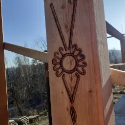 Drewniany słup z wyżłobionym ornamentem w kształcie słońca i strzałek, element konstrukcji domu w trakcie budowy, widok na zewnątrz z paletami i drzewami w tle.