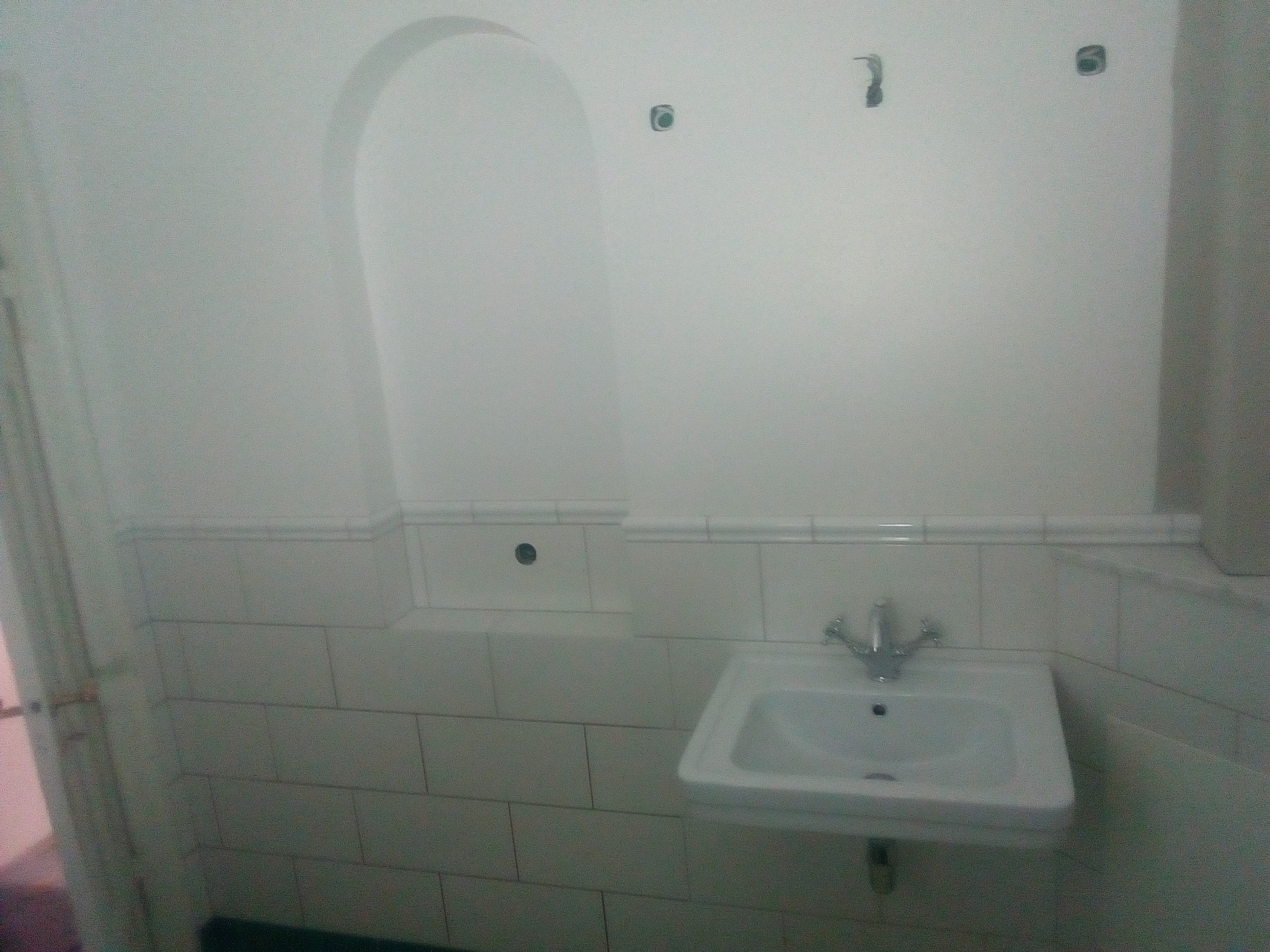 Biała umywalka z chromowaną baterią na tle białych płytek i ściany z wnęką w łazience w trakcie remontu, widoczne otwory na akcesoria.