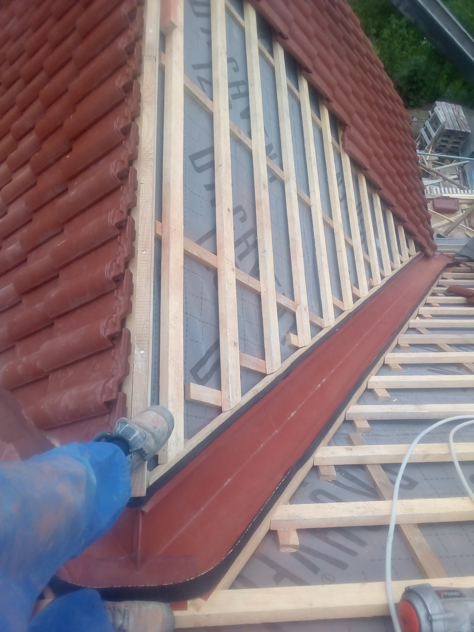 Montaż czerwonej dachówki ceramicznej na drewnianej więźbie dachowej z widoczną folią paroizolacyjną i fragmentem rynny, z butem pracownika w dolnym lewym rogu.