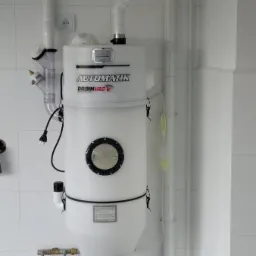 Biała jednostka centralna odkurzacza z napisem 'AUTOMATIK DRAIN VAC' zamontowana na kafelkowanej ścianie z widocznymi rurami instalacji i gniazdkiem elektrycznym.