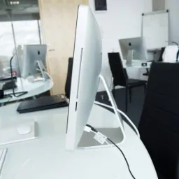 Nowoczesne biuro z iMacami na okrągłych stołach, widok z boku na monitor z podłączonymi kablami. Minimalistyczny design, białe meble, czarne krzesła, jasne wnętrze.