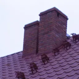 Dwa kominy z czerwonej cegły wystające z dachu pokrytego czerwoną dach&oacute;wką, z zamontowanymi stopniami kominiarskimi.