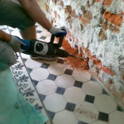 Pracownik w białych rękawicach używa młota udarowego do usuwania starego tynku z ceglanej ściany w pomieszczeniu z ozdobną, czarno-białą podłogą.