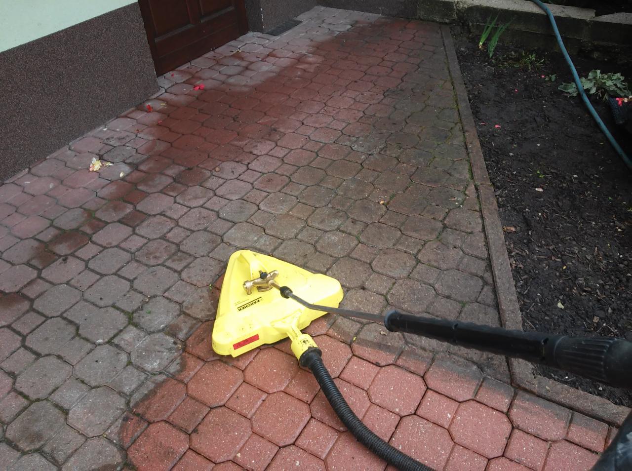 Czyszczenie kostki brukowej przed domem za pomocą myjki ciśnieniowej Karcher z żółtą przystawką do powierzchni płaskich, widoczne mokre i czyste fragmenty kostki.