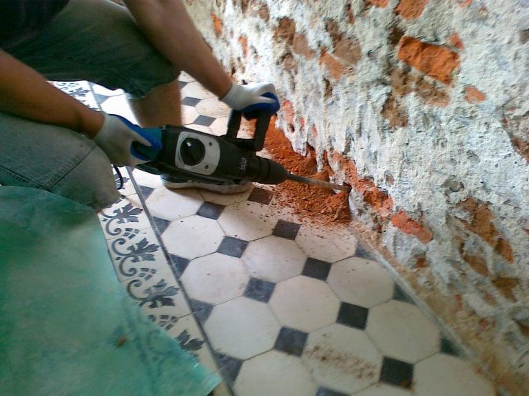 Pracownik w białych rękawicach używa młota udarowego do usuwania starego tynku z ceglanej ściany w pomieszczeniu z ozdobną, czarno-białą podłogą.