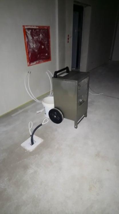 Metalowa osuszacz powietrza na kółkach, białe węże i kabel wystający z podłogi w surowym, szarym pomieszczeniu w trakcie prac wykończeniowych.