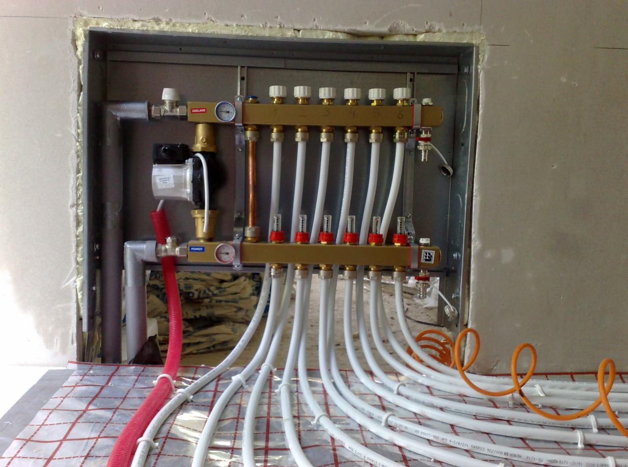 Rozdzielacz ogrzewania podłogowego z białymi rurami na folii grzewczej, widoczne manometry i zawory, instalacja w trakcie montażu w surowym pomieszczeniu.