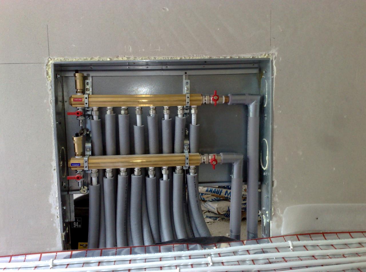 Rozdzielacz ogrzewania podłogowego w metalowej obudowie, z widocznymi rurami w szarej izolacji, zaworami z czerwonymi pokrętłami i fragmentem instalacji podłogowej w tle.