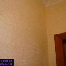 Veldecor - Ściana wykończona dekoracyjnym tynkiem trawertynowym w odcieniach beżu, widoczny fragment drewnianej framugi drzwi i listwa sufitowa.