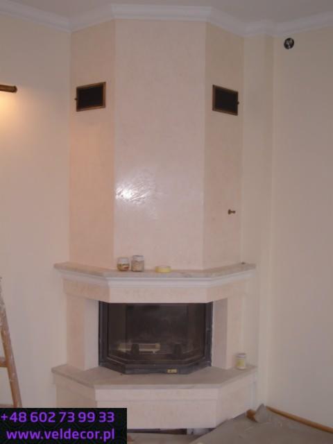 Wykończony kominek narożny ze stiukiem na ścianie, widoczne kratki wentylacyjne, obok drabina malarska.