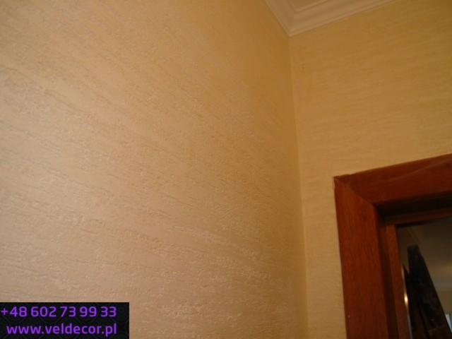 Ściana wykończona dekoracyjnym tynkiem trawertynowym w odcieniach beżu, widoczny fragment drewnianej framugi drzwi i listwa sufitowa.