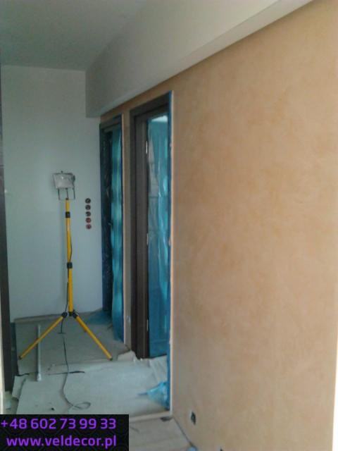 Ściana z efektem stiuku w odcieniach beżu, wykonana przez firmę z Lidzbarka, widoczna lampa budowlana i zabezpieczone folią drzwi.