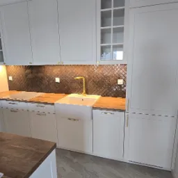 Biała kuchnia z zabudową meblową, złotą armaturą i mozaiką na ścianie, widoczne akcesoria montażowe na blacie.