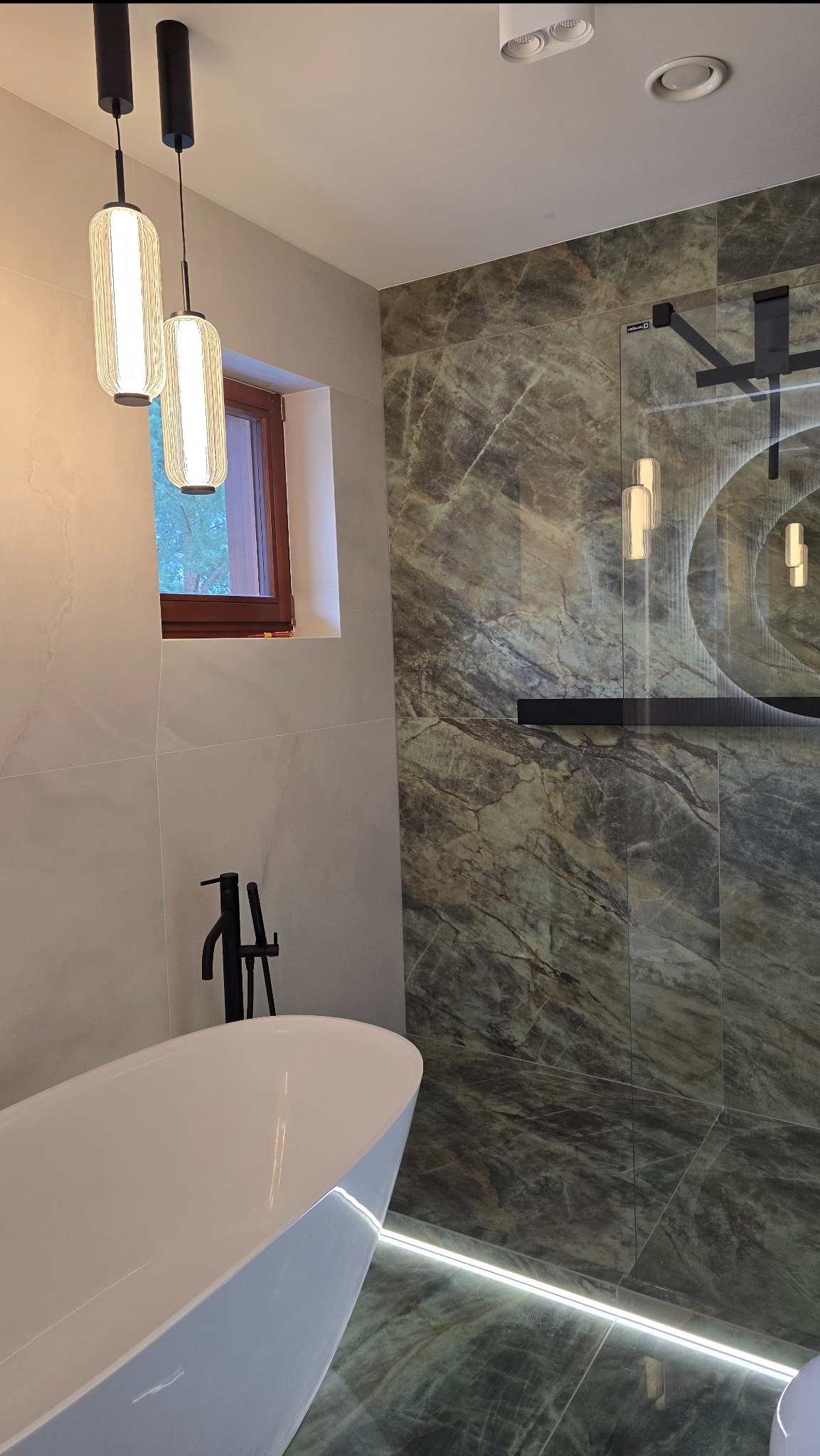 Nowoczesna łazienka z wolnostojącą białą wanną, czarną armaturą i marmurowymi płytkami z podświetleniem LED wzdłuż podłogi.