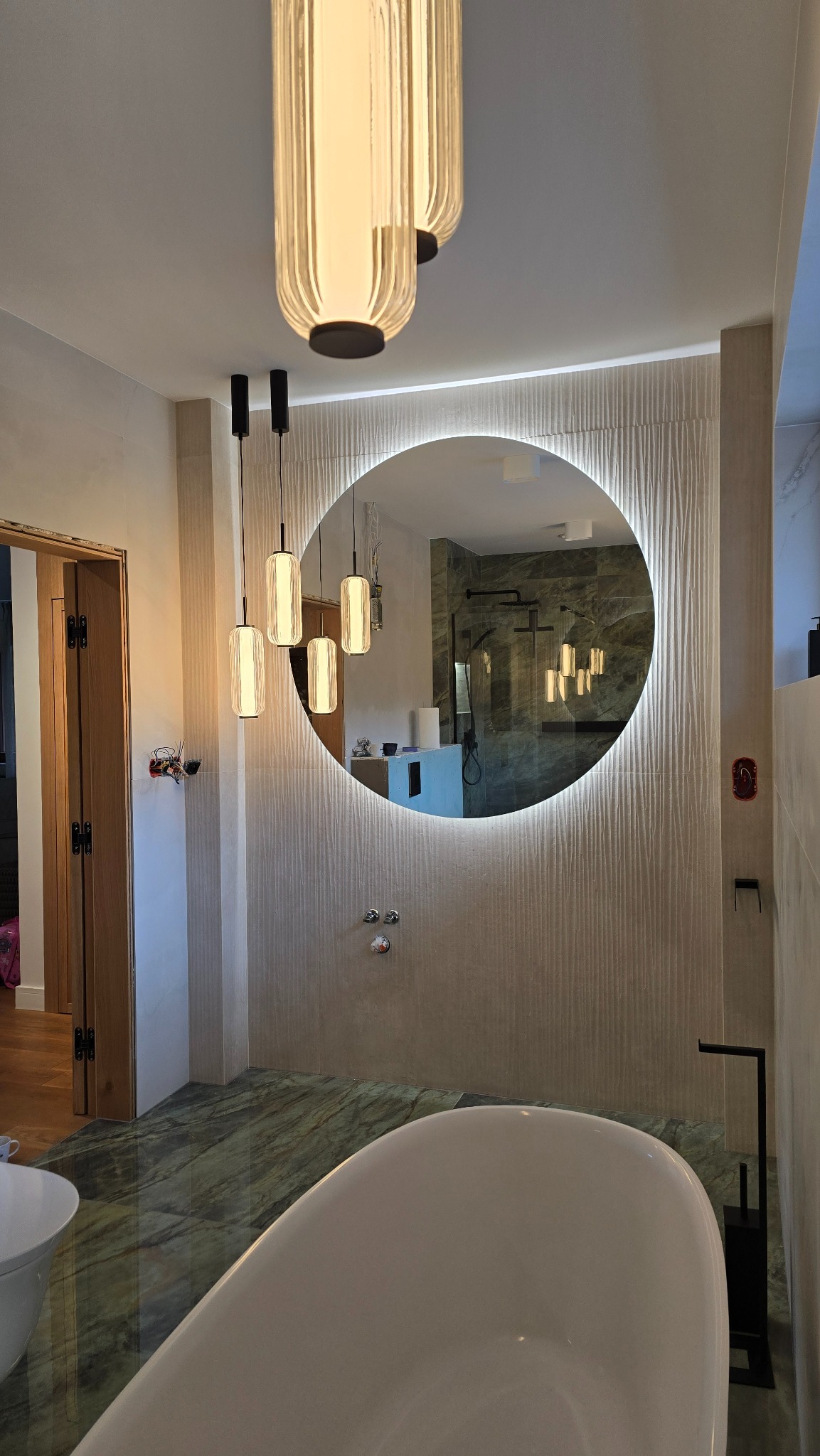 Nowoczesna łazienka z wolnostojącą wanną, okrągłym lustrem z podświetleniem LED, designerskimi lampami wiszącymi i marmurową podłogą.