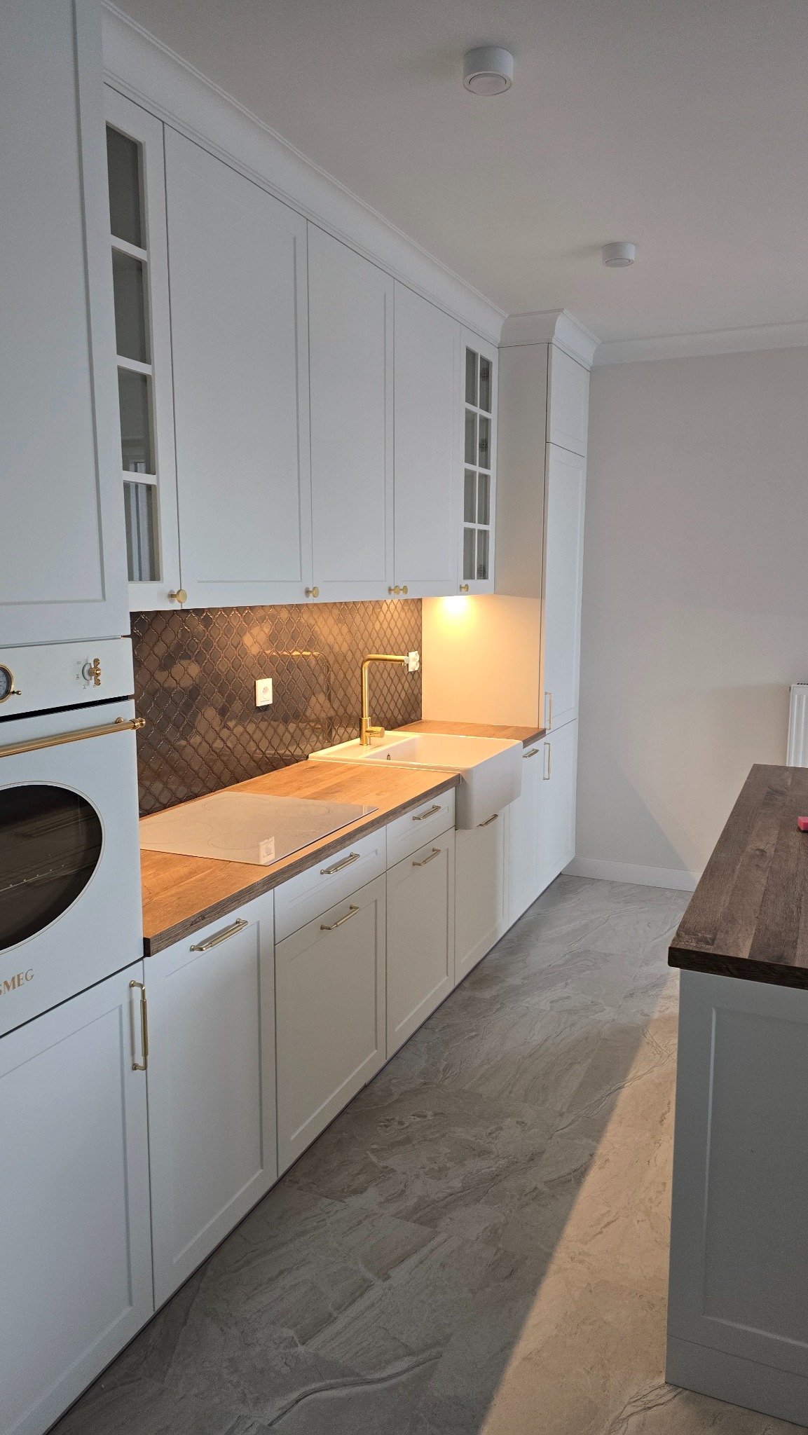 Biała kuchnia w stylu klasycznym z drewnianym blatem, złotą armaturą i mozaiką nad blatem roboczym, oświetlona punktowo.