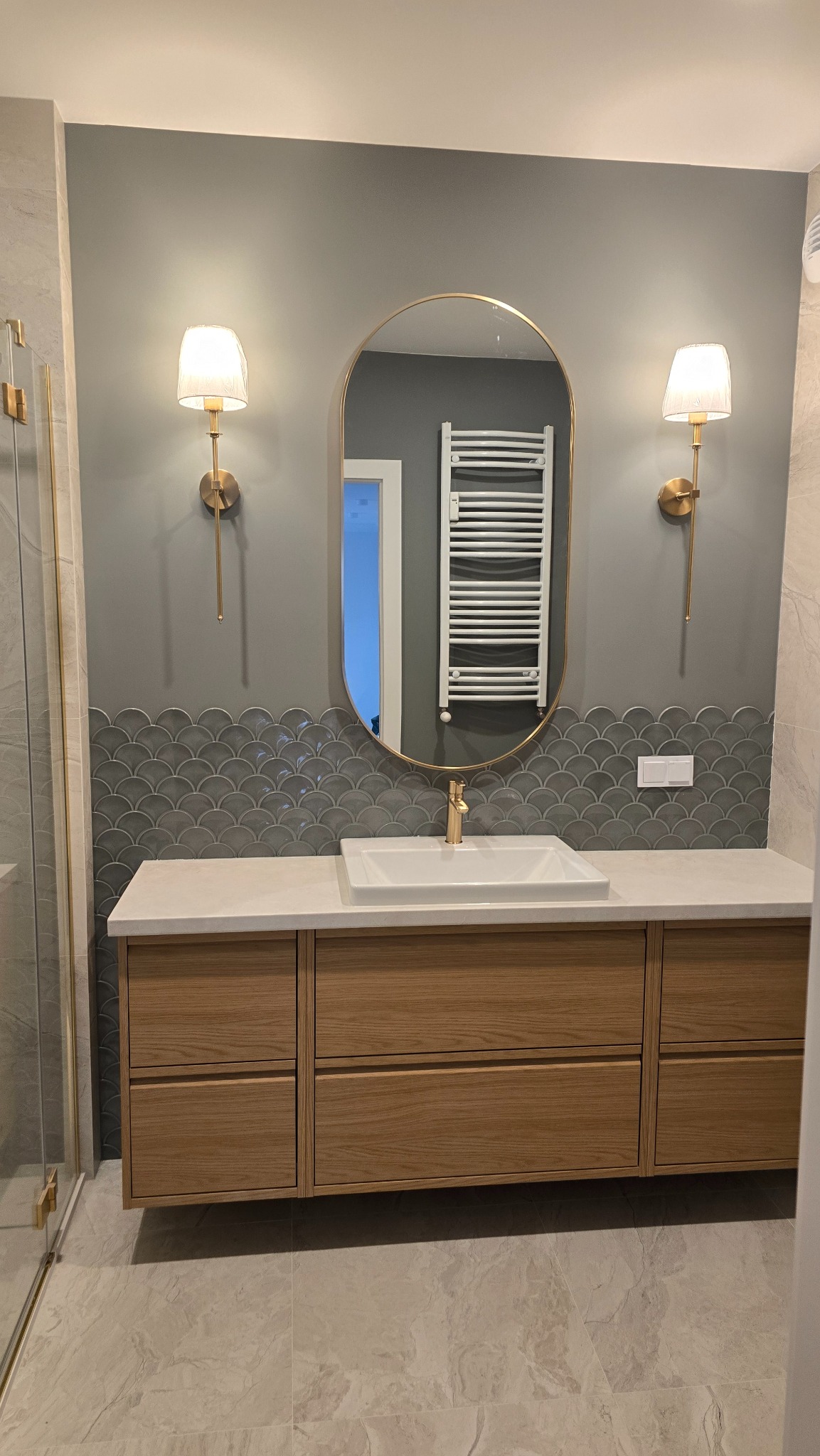 Nowoczesna łazienka z drewnianą szafką pod umywalką, owalnym lustrem w złotej ramie i dekoracyjnymi płytkami w kształcie rybiej łuski na ścianie.