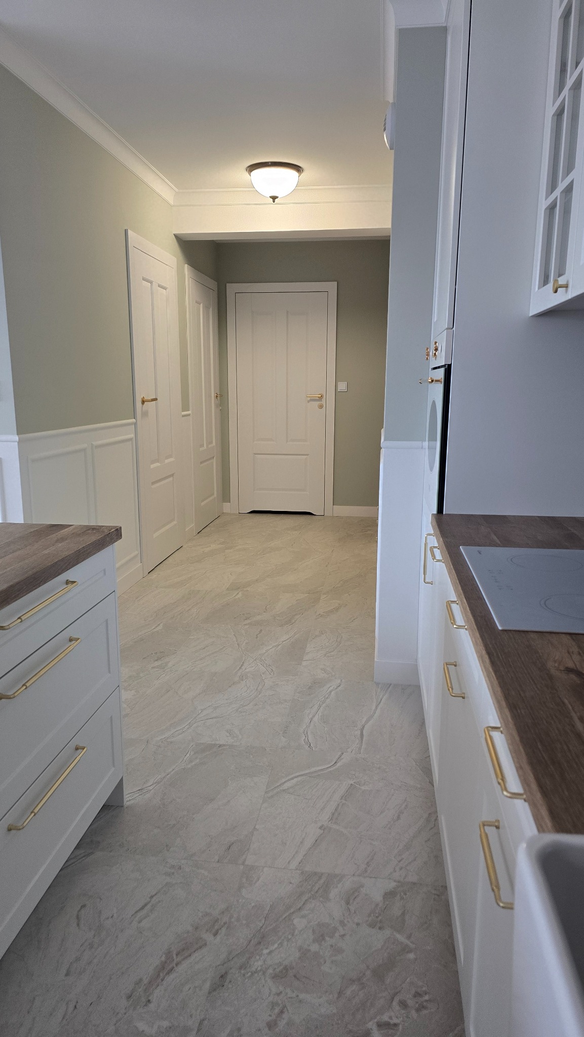 Jasne, marmurkowe płytki podłogowe w korytarzu z widokiem na białe drzwi i fragment zabudowy kuchennej z drewnianym blatem i złotymi uchwytami.