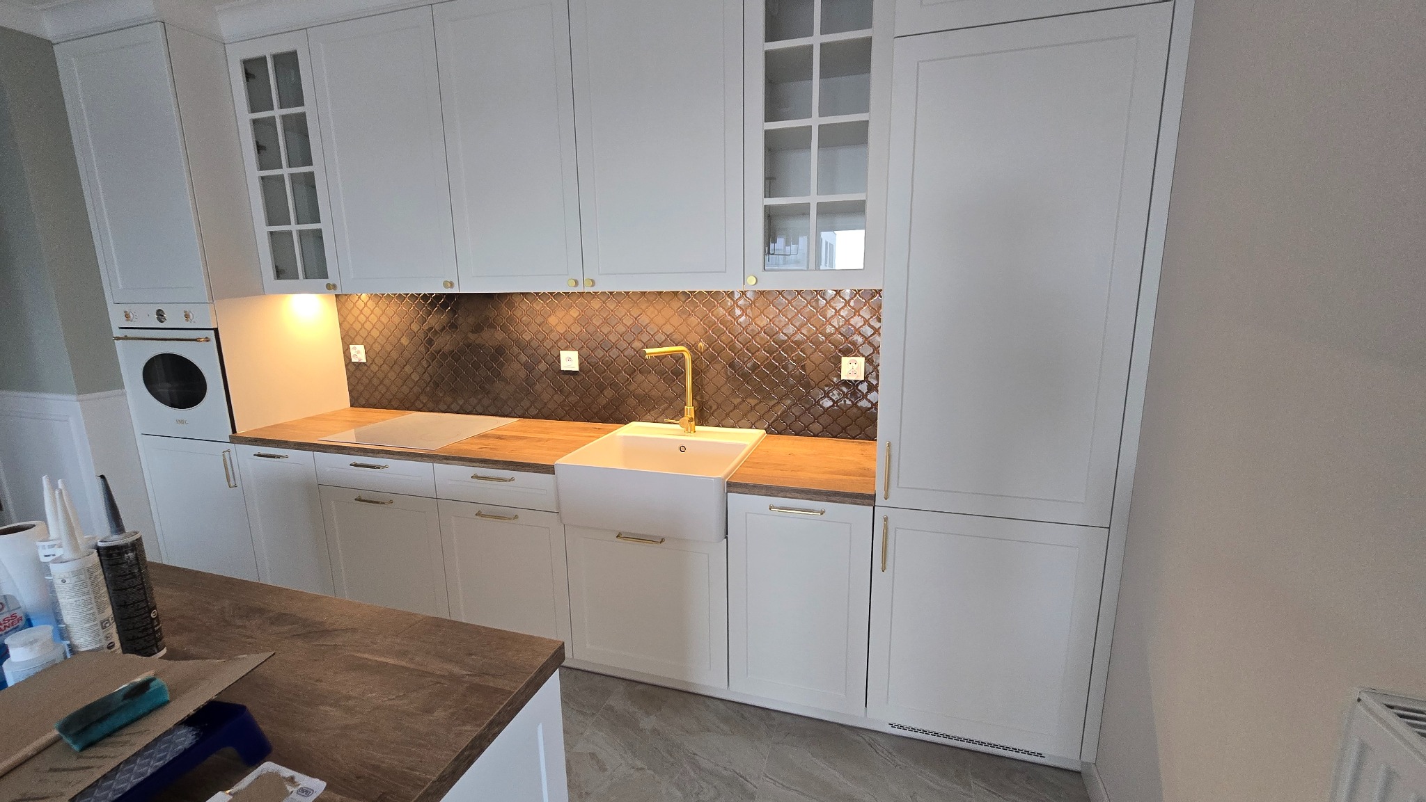 Biała kuchnia z zabudową meblową, złotą armaturą i mozaiką na ścianie, widoczne akcesoria montażowe na blacie.