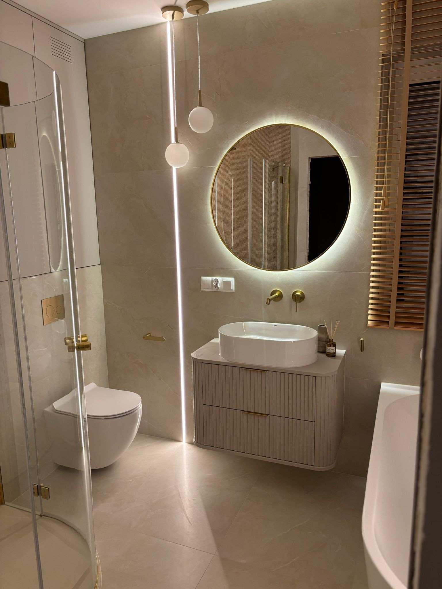 Elegancka łazienka w beżach z okrągłym lustrem podświetlanym LED, wiszącą toaletą i kabiną prysznicową ze złotymi detalami. Nowoczesny design z subtelnym oświetleniem.