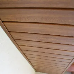 Wykończona podbitka dachowa z brązowych paneli imitujących drewno, widok od dołu, z fragmentem ściany o białej, chropowatej fakturze.